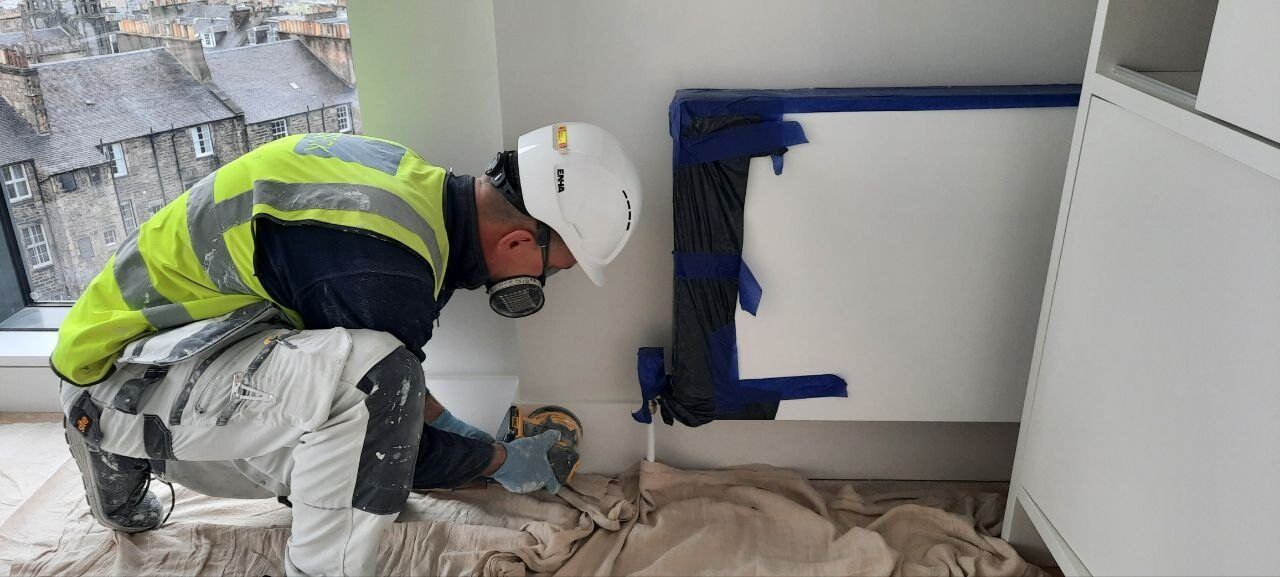 Vitalii in a hard hat and mask sanding beside a radiator on dust sheets